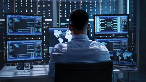System Security Specialist Working at System Control Center. Room is Full of Screens Displaying Various Information.