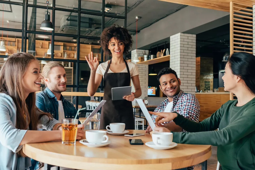  A smiling waitress with an apron and tablet interacts with a group of customers sitting at a table in a restaurant.